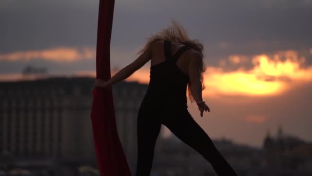 Sylwetka tancerki pokazuje swoją elastyczność i rozdziela się czerwonym powietrznym jedwabiem na tle pejzażu miasta podczas dramatycznego nieba w zwolnionym tempie. Pojęcie namiętności, pożądania i przyciągania — Wideo stockowe