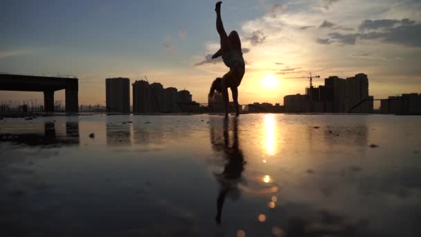 Flexibilní ženský gymnastka dělá handstand a kalisthenic s odrazem ve vodě na městském pozadí během dramatického západu slunce ve zpomaleném filmu — Stock video