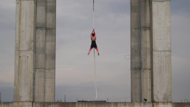 Сильный и мускулистый циркач, выступающий на воздушной веревке под открытым небом между двумя бетонными колоннами на фоне неба, Концепция мужества, контроля и выносливости — стоковое видео