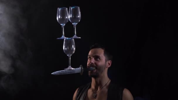 El hombre sostiene el cuchillo en la boca y mantener en equilibrio las gafas de vino en él. concentración — Vídeos de Stock
