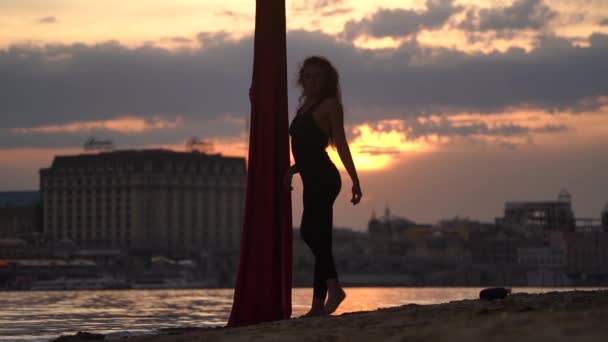 女性ダンサーのシルエットは、ゆっくりとした動きの中で劇的な空の間に背景に赤い空のシルクで彼女の柔軟性と分割を示しています。健康的なライフスタイルの概念. — ストック動画