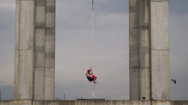 Sterke en gespierde circusartiest die optreedt op het touw buiten tussen twee betonnen zuilen op de achtergrond van de lucht, concept van kracht, risico en gevaar — Stockvideo