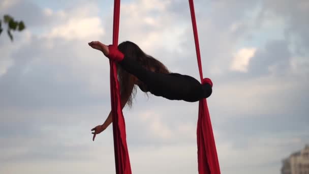 Acrobata fêmea mostrando sua flexibilidade e se divide com seda aérea vermelha no fundo do céu é câmera lenta. Conceito de graça, movimento e beleza — Vídeo de Stock
