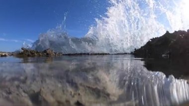 Bir deniz dalgası dalgası, volkanik kayalardan oluşan bir uçurumun kenarından süzülmesiyle dinginleşir, doğal havuzda kristal su dalgalanır ve arka planda mavi gökyüzü ile ağır çekimde kabarcıklar oluşur. 