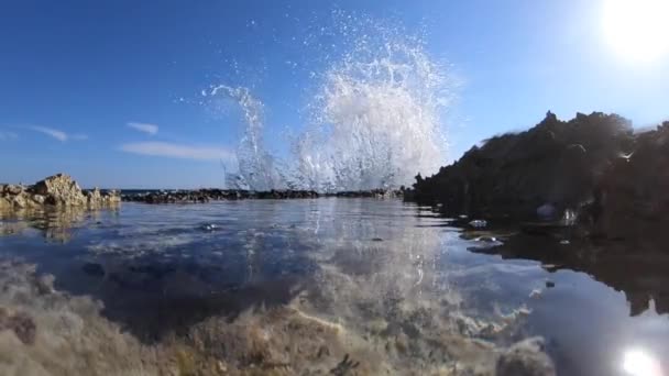 Гребень морской волны с белой пеной, которая ломается брызгами на скале вулканических пород в замедленной съемке с голубым небом на заднем плане — стоковое видео