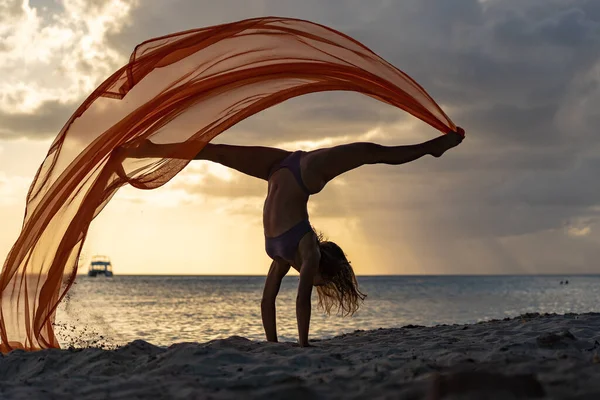 Silueta de mujer flexible haciendo handstand con seda durante la dramática puesta de sol con nubes tormentosas en el fondo del paisaje marino. Concepto de felicidad, libertad y despreocupación. — Foto de Stock