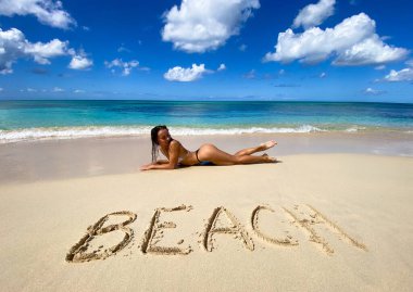 Seksi bikinili kadın kumdaki yazıyla birlikte tropikal bir plaja uzanıyor. Seyahat ve tatil kavramı