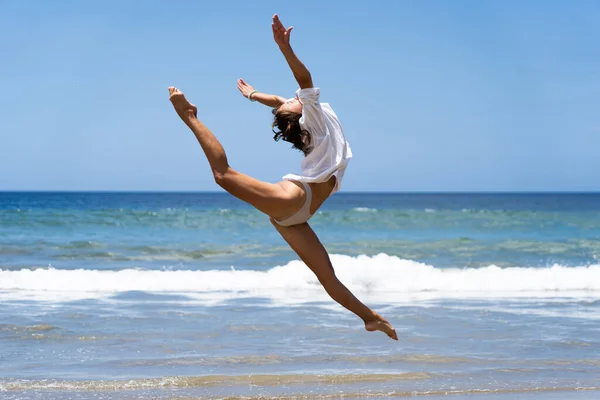 Joven bailarina alegre en traje de baño y camisa blanca mientras salta felizmente con el mar en el fondo. Concepto de despreocupación y felicidad — Foto de Stock