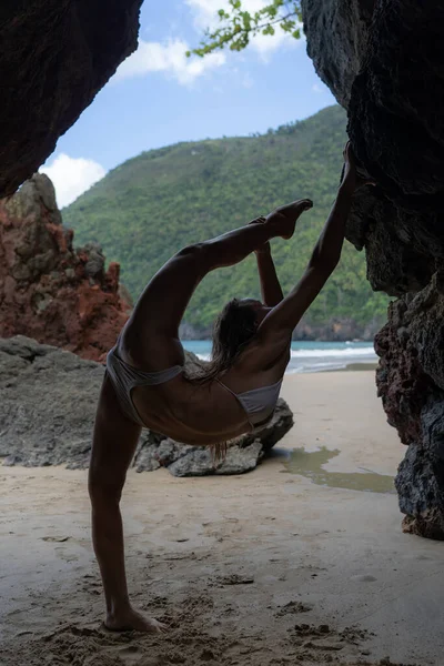 Mujer joven, en forma, sexy haciendo estiramiento en la cueva con la playa en el fondo. Concepto de yoga y flexibilidad — Foto de Stock
