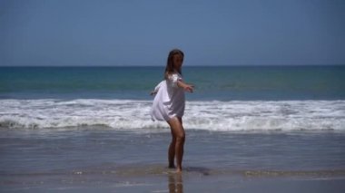 Mayo ve beyaz gömlekli neşeli genç bir kadın arka planda deniz varken ağır çekimde mutlu bir şekilde dans ediyor. Tatil ve seyahat kavramı 