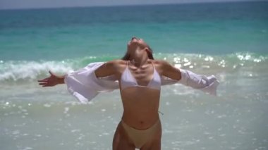 Mayo ve beyaz gömlekli neşeli genç bir kadın arka planda deniz varken ağır çekimde mutlu bir şekilde dans ediyor. Tatil ve seyahat kavramı 