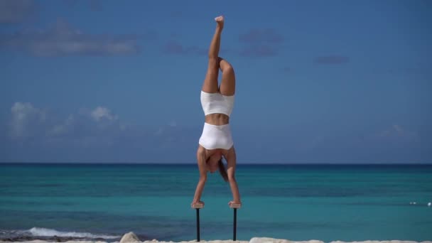 Женщина в белом делает стойку на руках на пляже с морем. Здоровый образ жизни и уход за телом. — стоковое видео