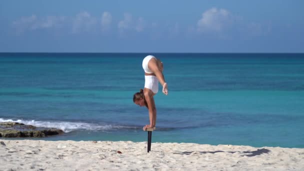 Kvinna klädd i vitt gör handstativ på stranden med havet. Individualitet, enastående och originalitet. — Stockvideo