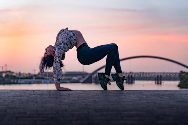 Yoga yapan esnek kadının silueti dramatik günbatımında ve şehir manzarasında sırtını eğiyor. Sağlıklı yaşam tarzı ve sağlık hizmeti kavramı 