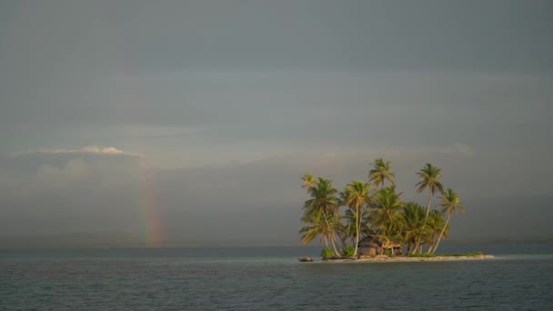 Denizde küçük tropikal, ıssız bir ada. Bulutlu gökyüzünde palmiye ağaçları ve gökkuşağı var. Tatil ve seyahat kavramı — Stok video