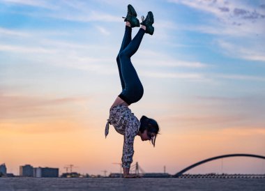 Esnek kadın silueti dramatik günbatımı ve şehir manzarasında amuda kalkıyor. İrade, motivasyon ve sağlıklı yaşam tarzı kavramı