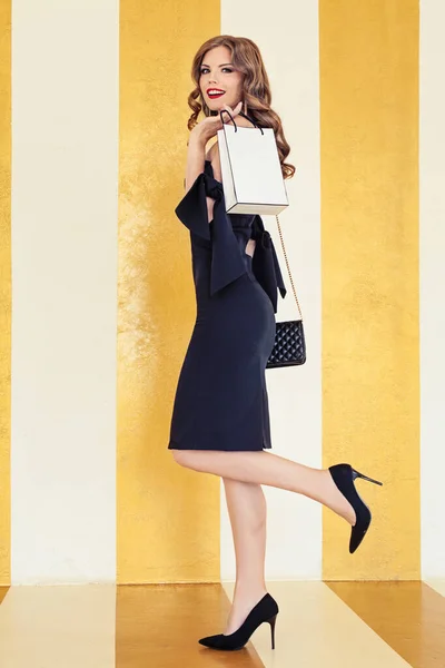 グラマラスな女性で黒カクテルドレス保持ショッピングバッグ上の金の背景 — ストック写真