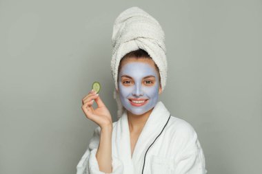 Beyaz arka planda salatalıkla nemlendirici kozmetik maske takan gülümseyen kadın spa modeli. Beyaz bornoz ve havlu giymiş bir kadın. Yüz bakımı ve cilt bakımı kavramı