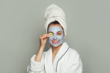 Gülümseyen genç bir kadın, yeşil salatalıklı nemlendirici kozmetik maske takıyor. Yüz bakımı ve cilt bakımı kavramı