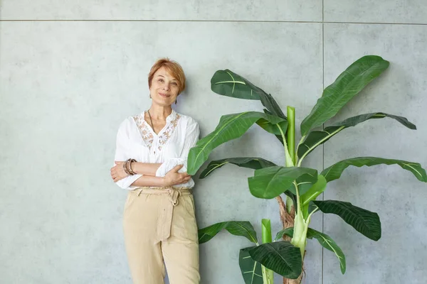 健康なシニア女性笑顔と緑の葉植物と灰色の背景に交差腕で立って — ストック写真