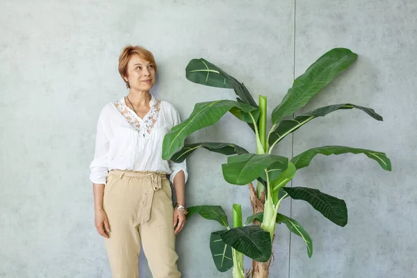 灰色の背景に緑の植物の近くに立つ白いシャツのシニア女性の肖像画 — ストック写真