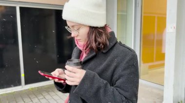 Cep telefonlu genç bir kadın, donmuş şehrin caddesinde sıcak bir içki içiyor.. 