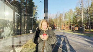 Güneşli bir günde otobüsü bekleyen cep telefonlu genç bir kadın.