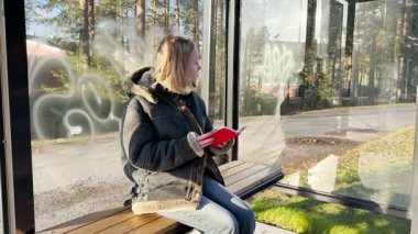 Kırmızı ders kitabı olan genç bir kadın küçük bir kasabada şehir otobüsünü bekliyor.