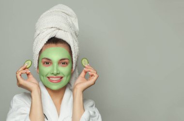 Yeşil organik kozmetik maskeli, elinde salatalık dilimleri olan mutlu bir kadın.