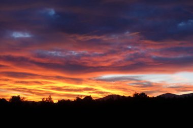 Karanlık ağaçlar ve tepeler üzerinde günbatımı renkleriyle bulutlu gökyüzü.