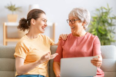 Genç ve yaşlı kadınlar dizüstü bilgisayar kullanıyorlar. Kız, anneye evde kalarak eğlenmesi için yardım ediyor..