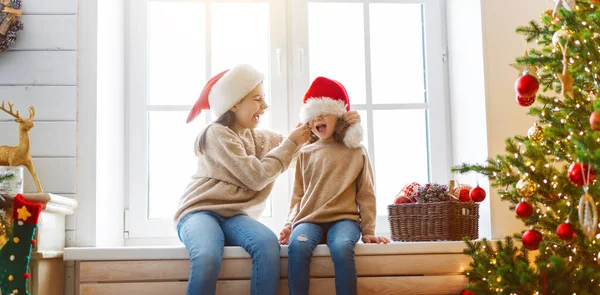 メリークリスマスとハッピーホリデー ウィンドウに座って陽気なかわいい子供たちの女の子 子供たちは朝木の近くで楽しんでいます — ストック写真