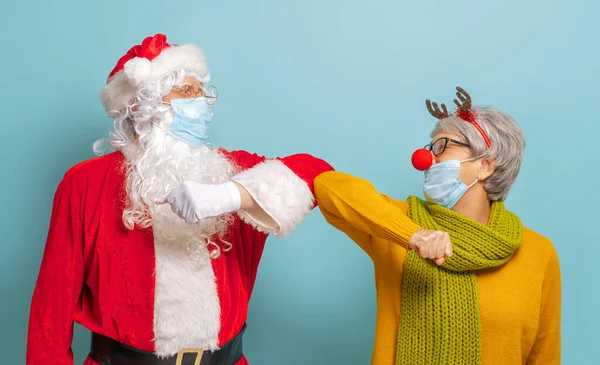 メリークリスマスとハッピーホリデー 明るい色の背景にサンタクロースとトナカイ Covid 2019の間にフェイスマスクを身に着けているカーニバル衣装の高齢者 — ストック写真