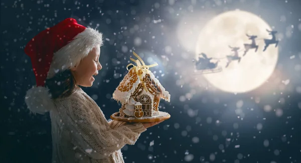 メリークリスマス ジンジャーブレッドハウスのかわいい子供 サンタクロースは月の空に対して彼のそりで飛んでいます 幸せな子供は休日をお楽しみください 暗い背景の女の子の肖像画 — ストック写真