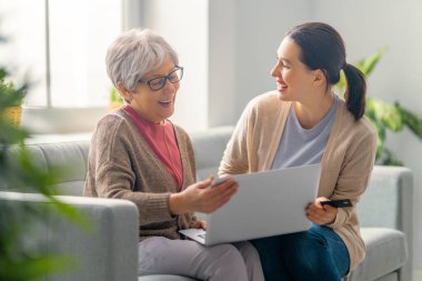 Genç ve yaşlı kadınlar dizüstü bilgisayar kullanıyorlar. Kız, anneye evde kalarak eğlenmesi için yardım ediyor..
