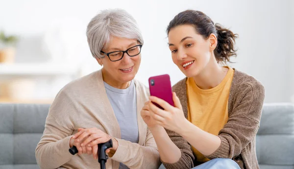 若い女性や年配の女性がスマートフォンを使っています 娘は母親が家にいるのを楽しんでるのを助けてる — ストック写真