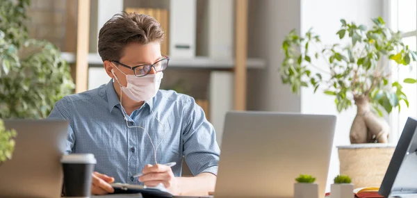 Young man in face mask is working on a laptop in office.