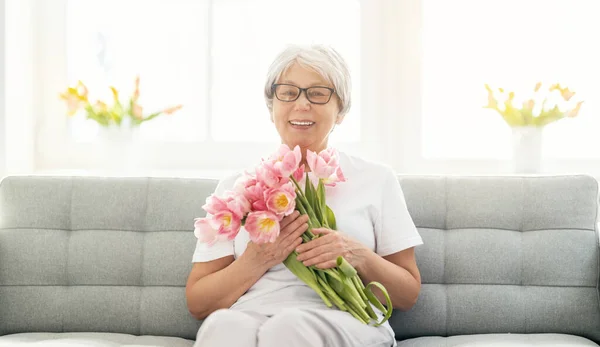 Красивая Пожилая Женщина Розовыми Цветами Тюльпаны Руках Сидя Диване Дома — стоковое фото