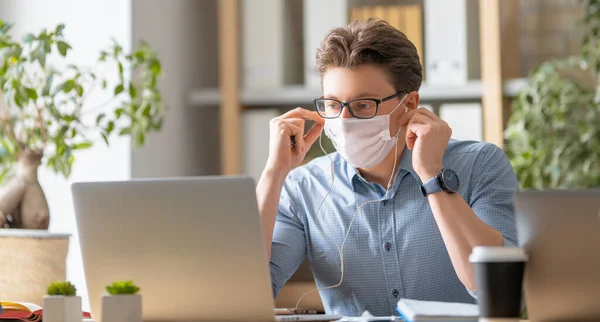 顔のマスクの若い男がオフィスのラップトップで働いています — ストック写真