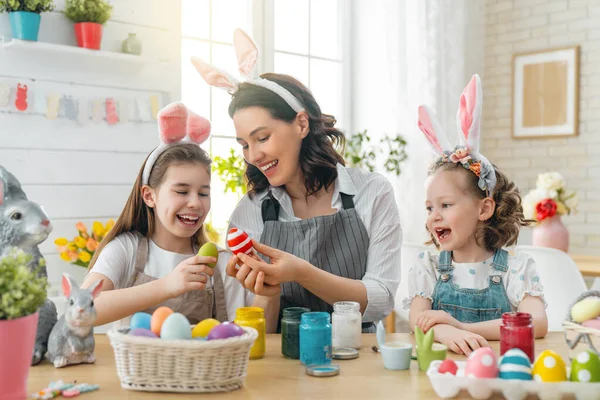 Happy holiday! A mother and her daughters are painting eggs. Family preparing for Easter. Cute little children girls are wearing bunny ears.