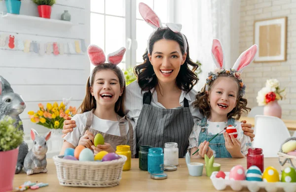 Happy holiday! A mother and her daughters are painting eggs. Family preparing for Easter. Cute little children girls are wearing bunny ears.