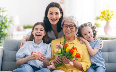 Anneler Günün kutlu olsun! Çocuk kızları ve anne, büyükannenin ona çiçek lalesi vermesini kutluyorlar. Büyükanne, anne ve kızlar gülümsüyor ve sarılıyorlar. Aile tatili ve birliktelik.