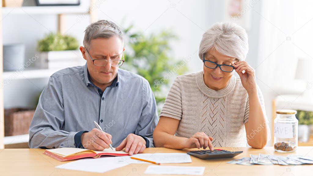 Elderly married couple sitting at the desk with a paper receipt in hands are calculating expenses, managing the family budget.