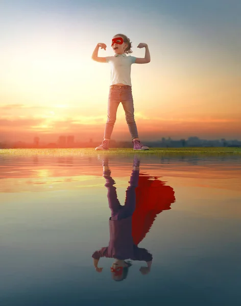 幼い子供がスーパーヒーローを演じて水面に映る姿を見ている 日没の空の背景に子供 ガールパワーコンセプト — ストック写真