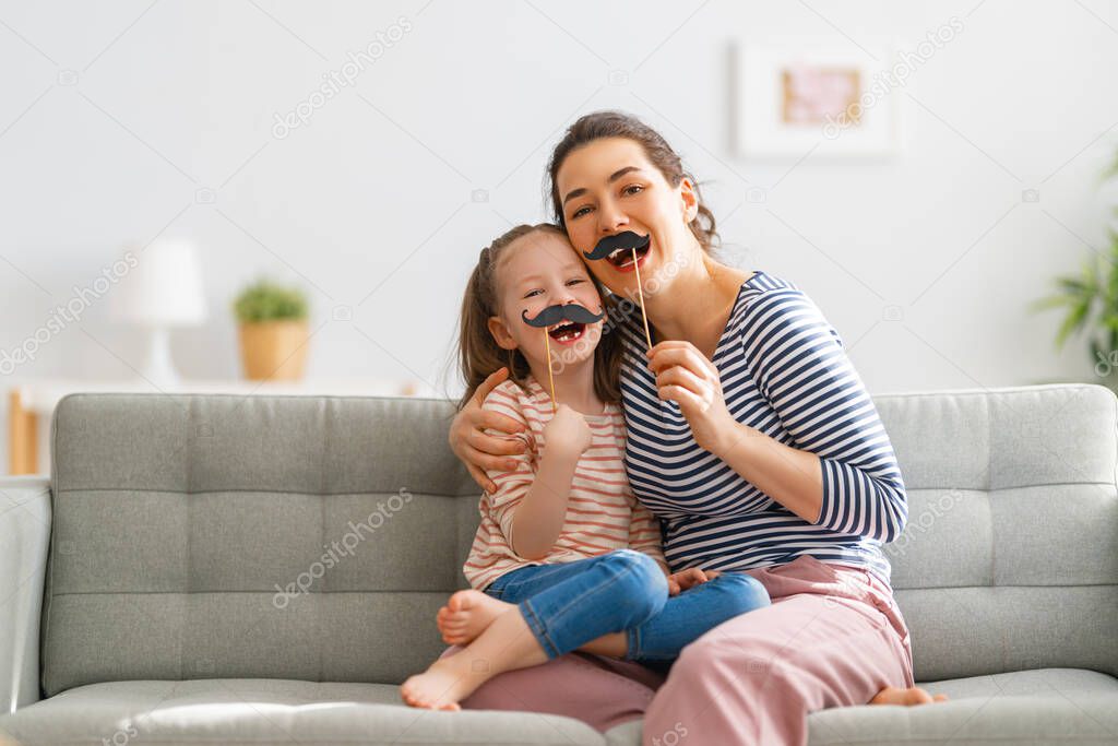 Mom and her child daughter are playing at home. Cute girl are holding paper mustache on stick. Family holiday and togetherness.