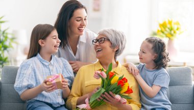 Anneler Günün kutlu olsun! Çocuk kızları ve anne, büyükannenin ona çiçek lalesi vermesini kutluyorlar. Büyükanne, anne ve kızlar gülümsüyor ve sarılıyorlar. Aile tatili ve birliktelik.