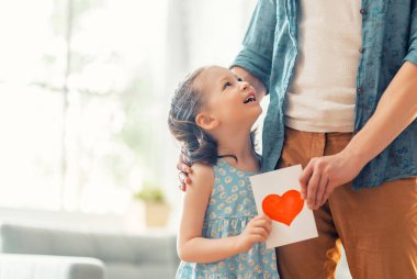 Çocuk kızı babasını tebrik ediyor ve ona kartpostal veriyor. Baba ve kız gülümsüyor ve sarılıyorlar. Aile tatili ve birliktelik.