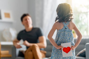 Çocuk kızı babasını tebrik ediyor ve ona kartpostal veriyor. Baba ve kız gülümsüyor ve sarılıyorlar. Aile tatili ve birliktelik.