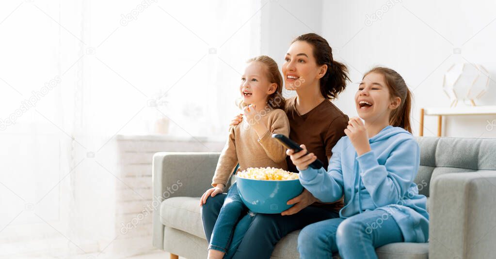 Happy family watching TV, movies with popcorn in the evening at home. Mother and daughters spending time together.