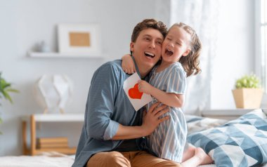 Çocuk kızı babasını tebrik ediyor ve ona kartpostal veriyor. Baba ve kız gülümsüyor ve sarılıyorlar. Aile tatili ve birliktelik.